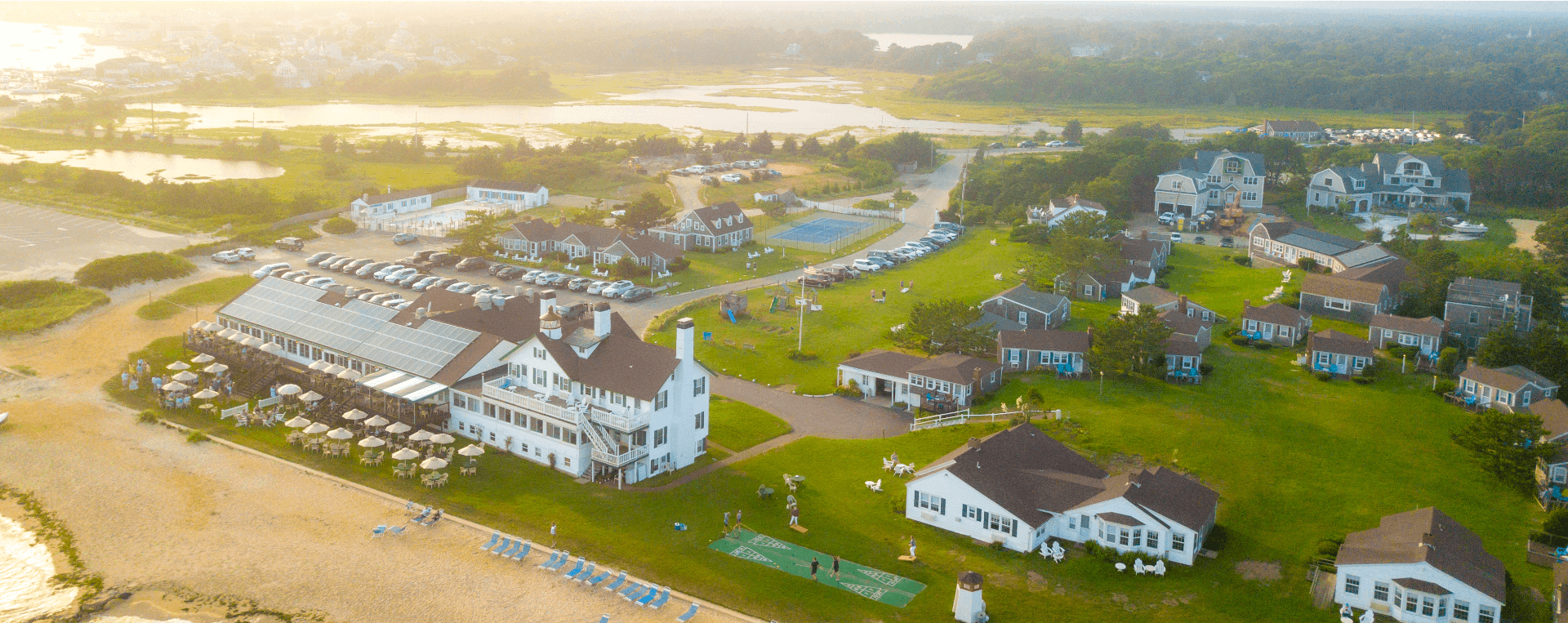 An aerial view of a large white hotel complex with brown roofs, surrounded by green grass and a beach.