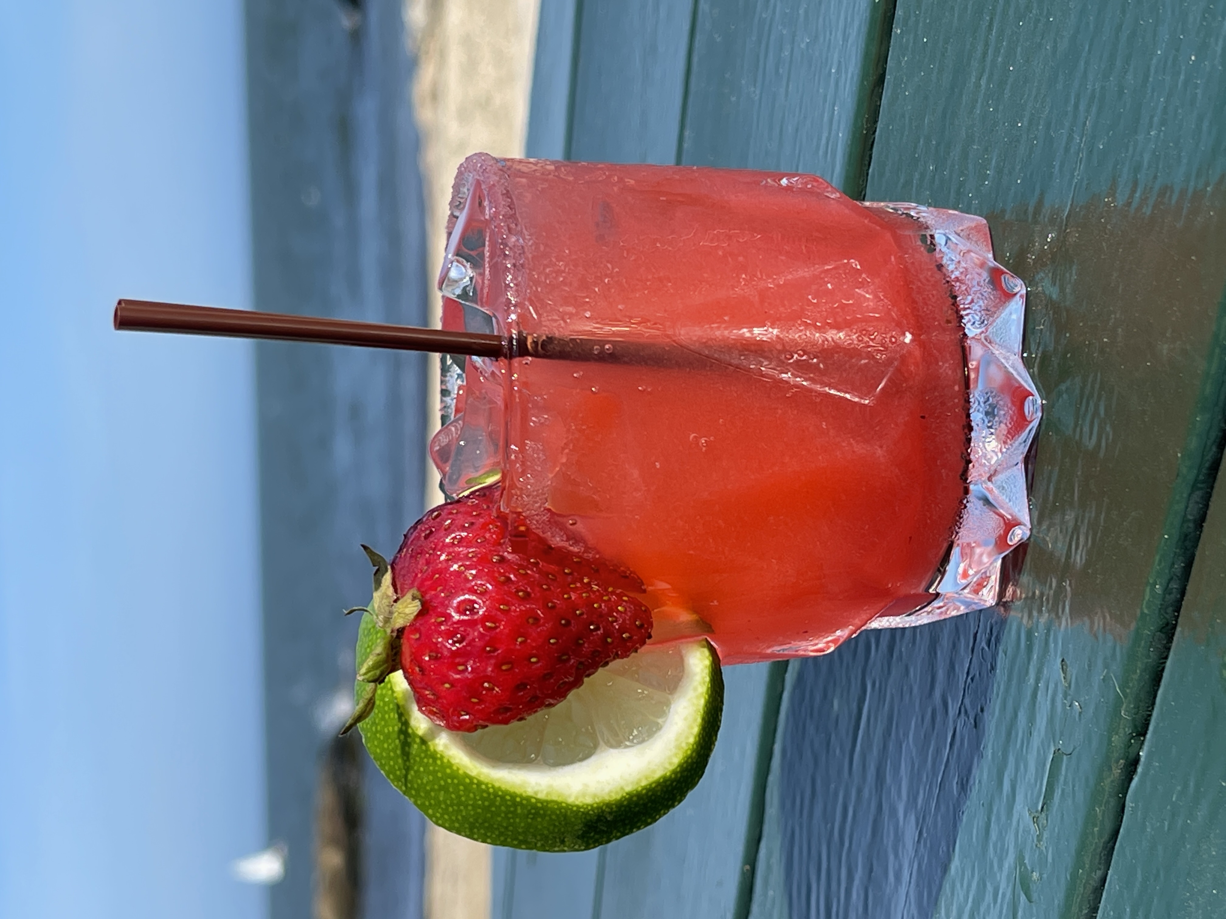 a pink cocktail with a strawberry and lime garnish on a wooden table with the ocean in the background